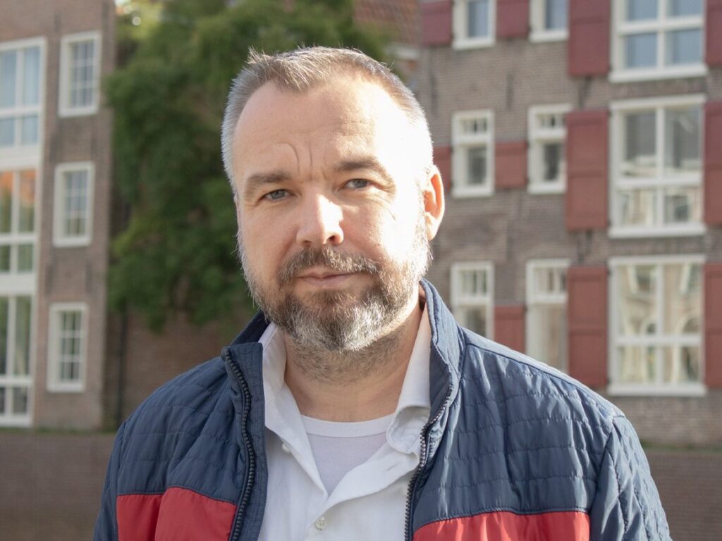 Portret van Leander Broere in jas, staand buiten bij gebouwen met raamluiken