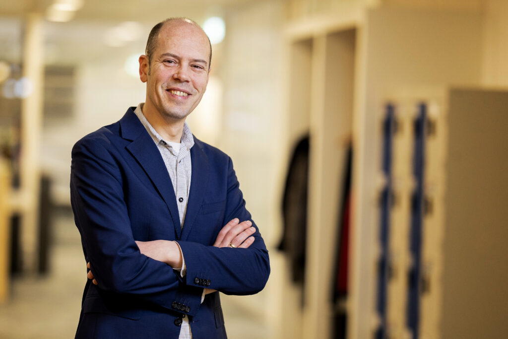 Portret van Nanco Dolman in blauw colbert, staand met armen over elkaar in kantooromgeving